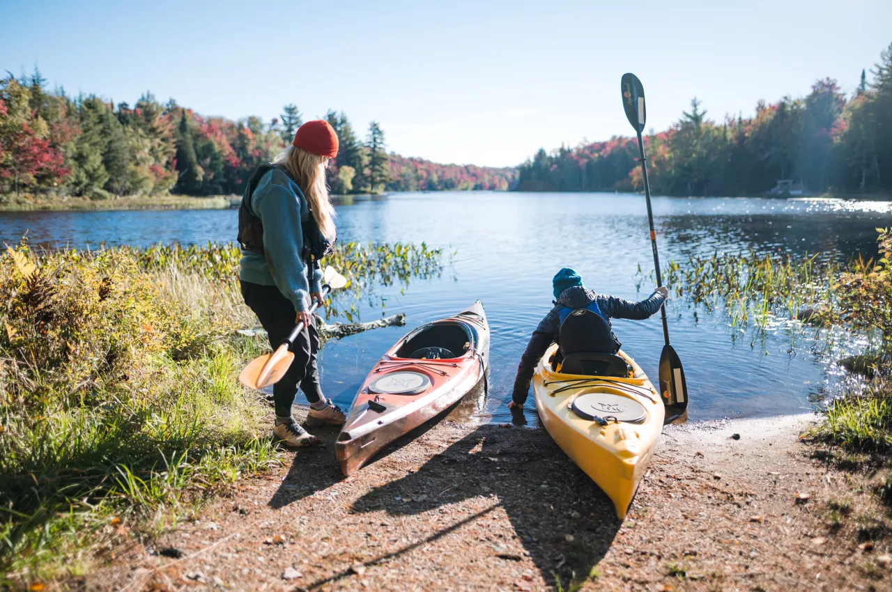 One person stands next to a kayak on the shore of an Adirondack lake, and a second person sits in an adjacent kayak