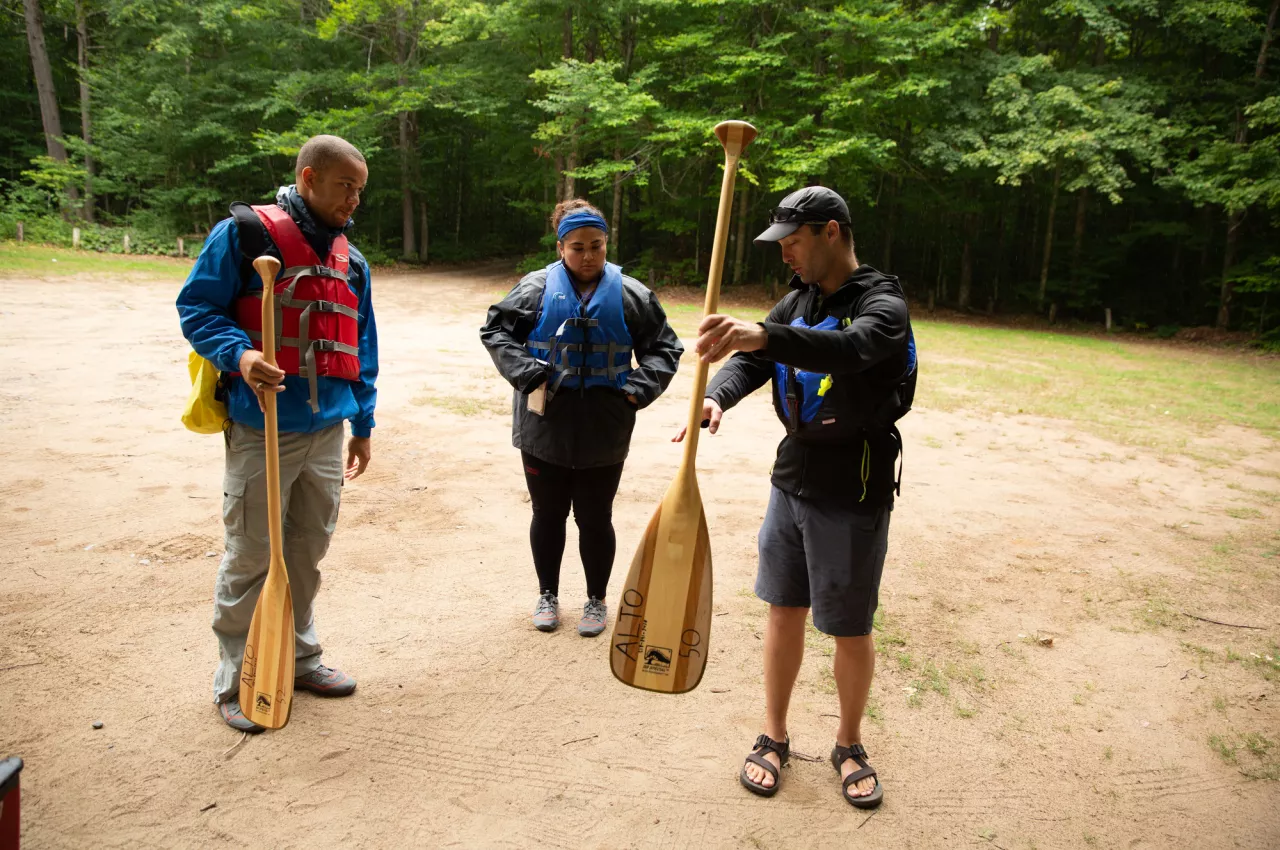 Three people stand near each other holding canoe paddles as one appears to explain paddling technique to the other two.