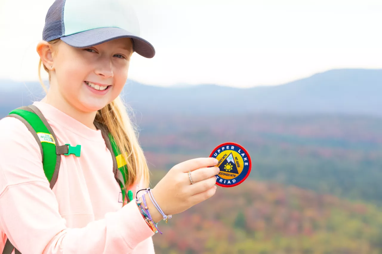 Girl holds up a Tupper Lake Triad Patch and smiles at the camera a the top of a mountain.