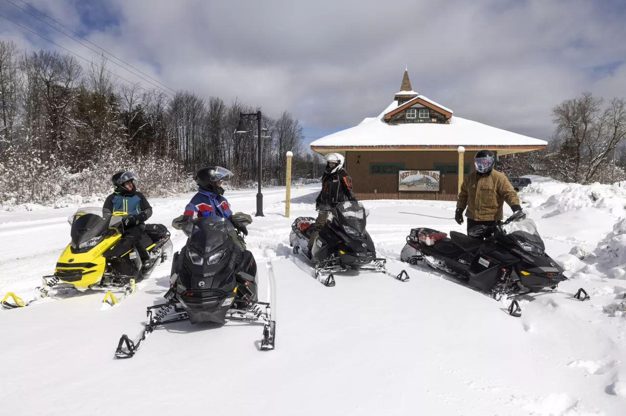 A group of four snowmobilers pause on their sleds in front of a snowy historic train station.