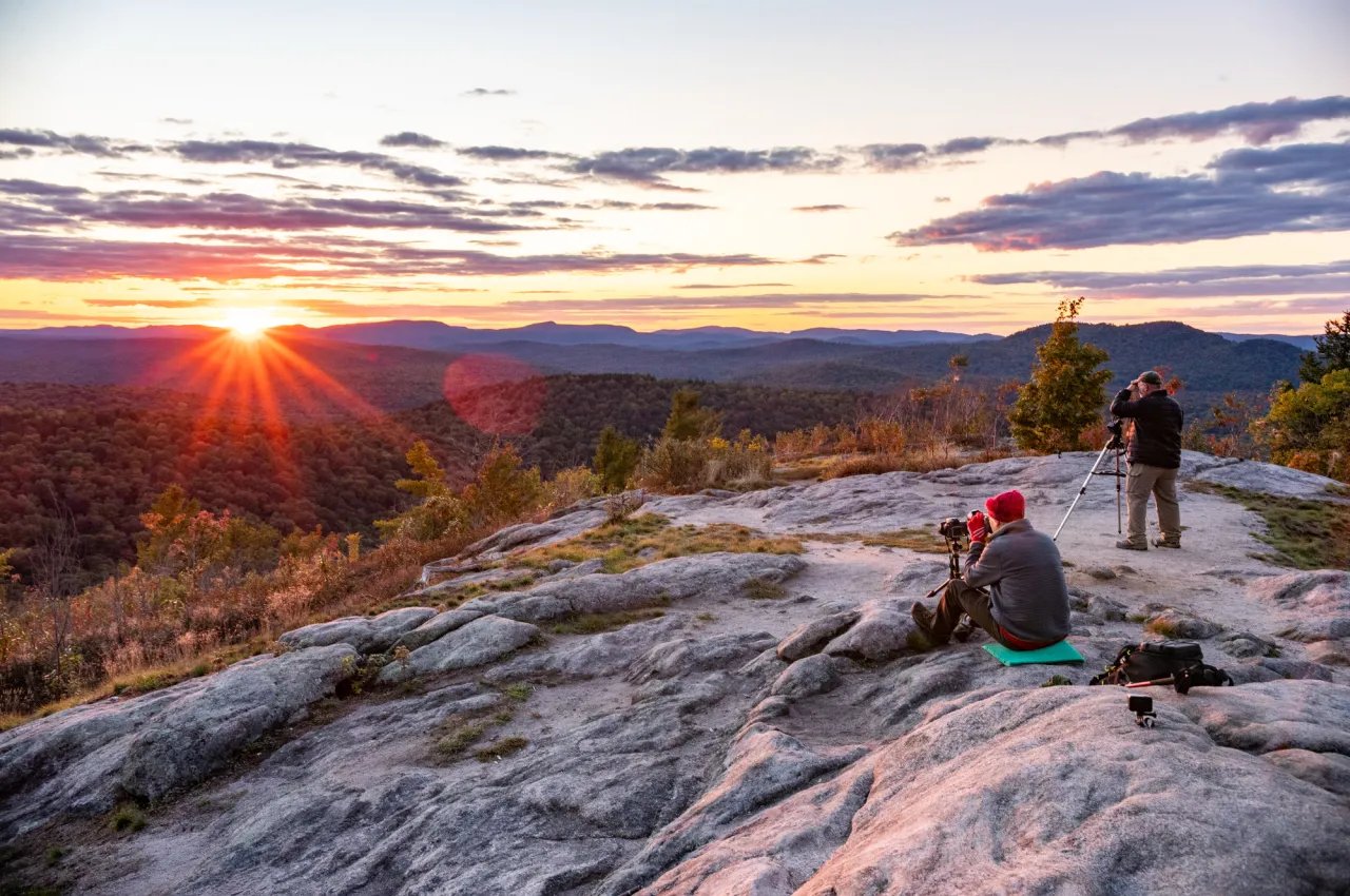 Two hikers in cool weather clothing photograph a sunset from a mountain summit.