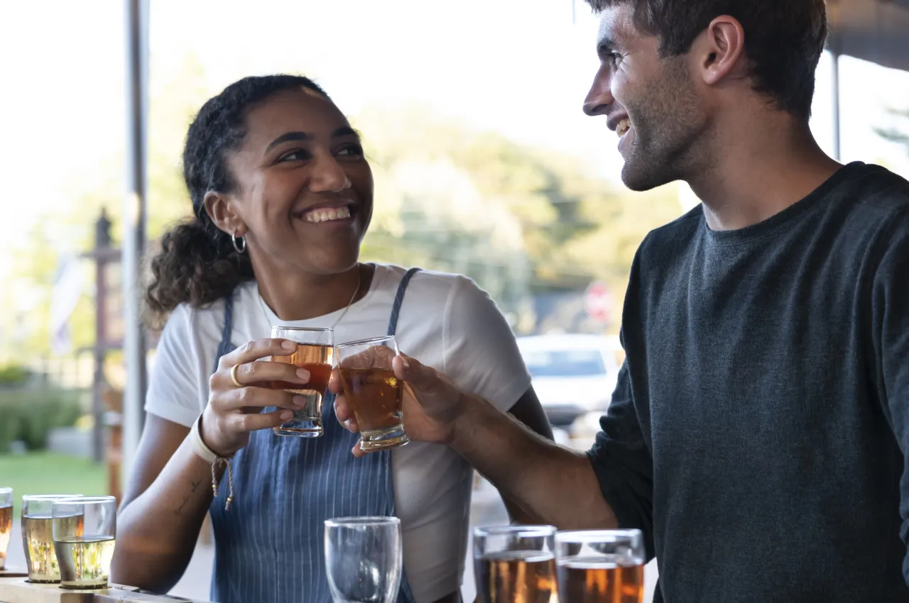 A woman and a man clink small glasses of beer while smiling at each other.