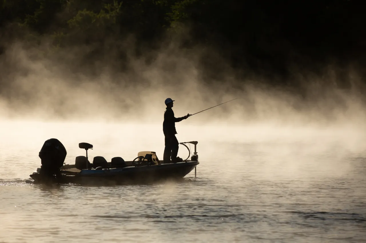 A man on a fishing boat is silhouetted against a misty lake.