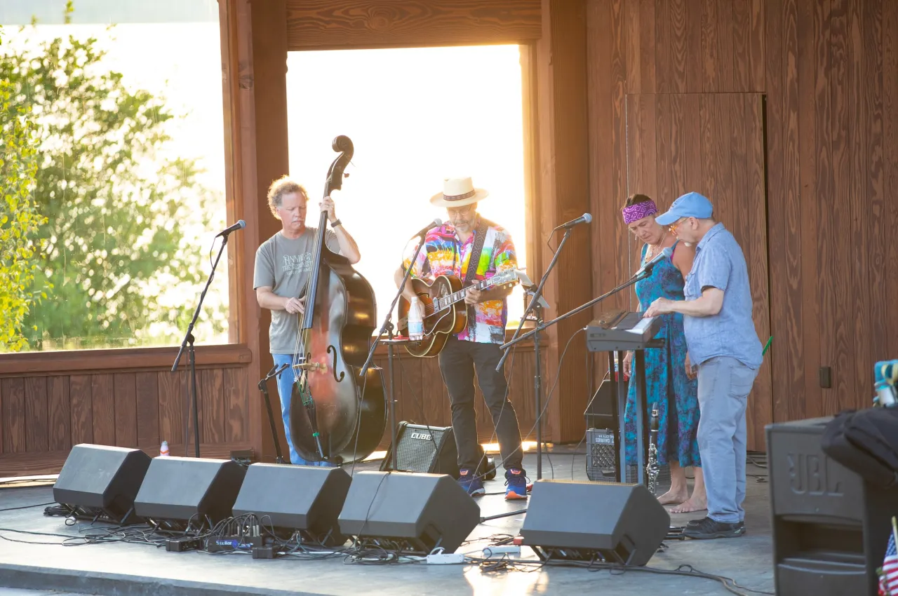 Four adults perform live music on a sunny outdoor wood bandshell.