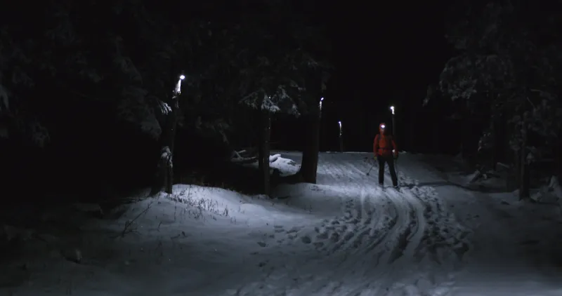 A man with a headlamp skis downhill past lights at night