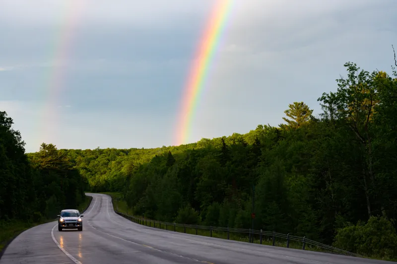 a car drives on a wet road with a rainbow in the sky behind it.