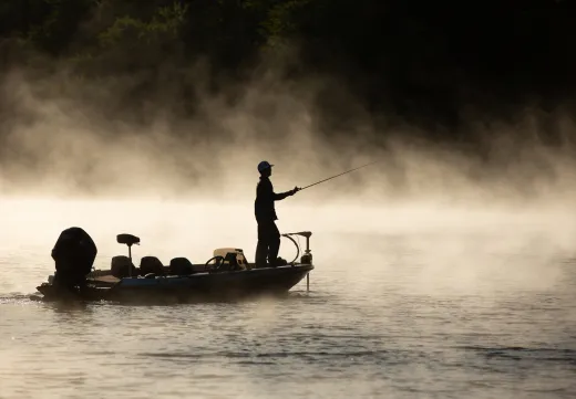 A man fishes off a boat amid sunrise mist