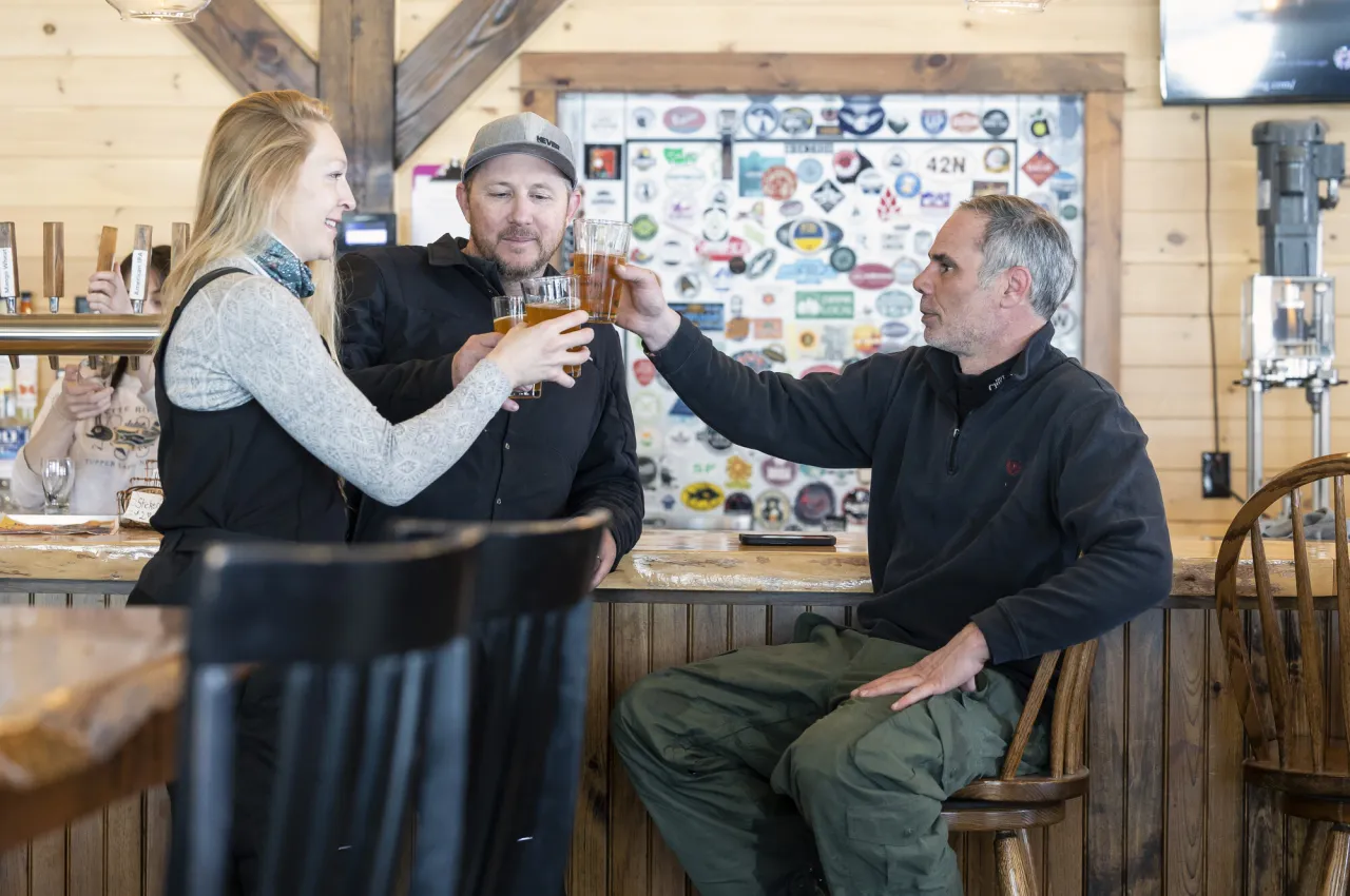 Three adults clink drinks at a cozy, rustic bar in winter.
