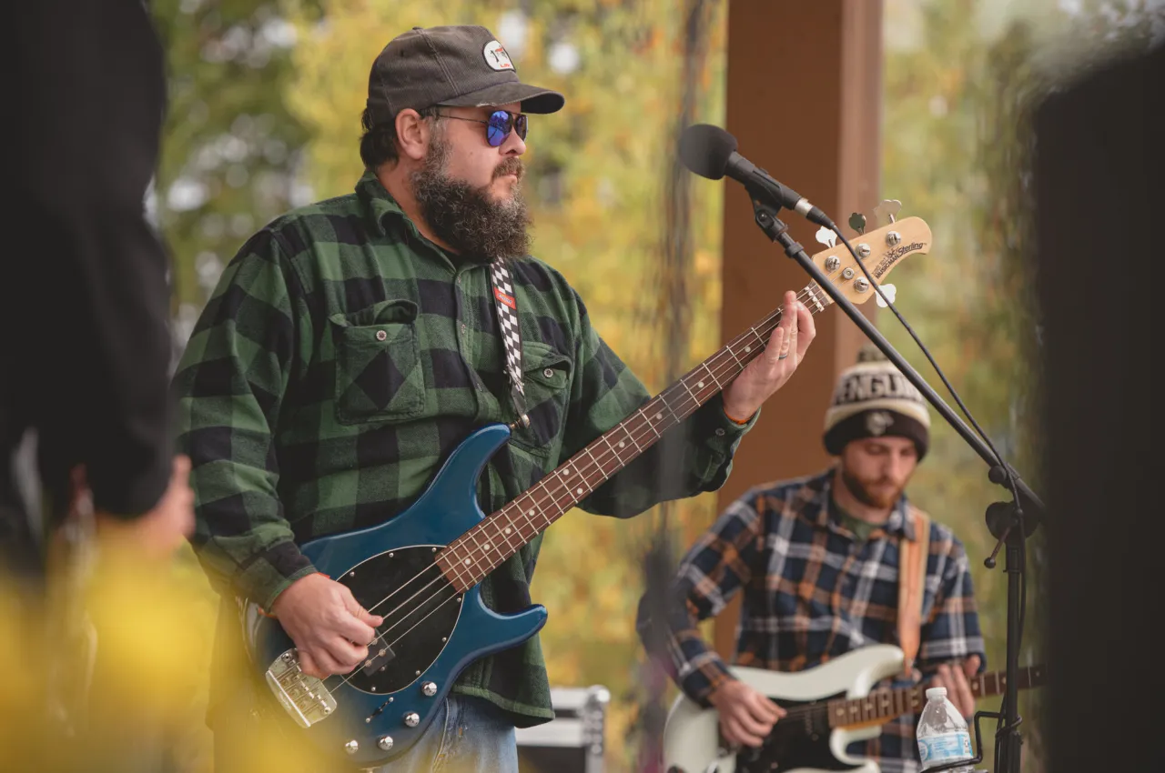 A bearded man wearing sunglasses and a baseball cap plays bass on an outdoor stage.