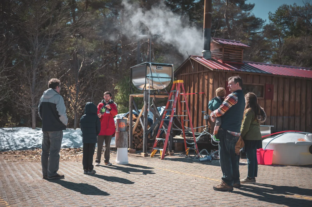 A small group of adults and children listen to a guide talk outside a maple sugar shack.