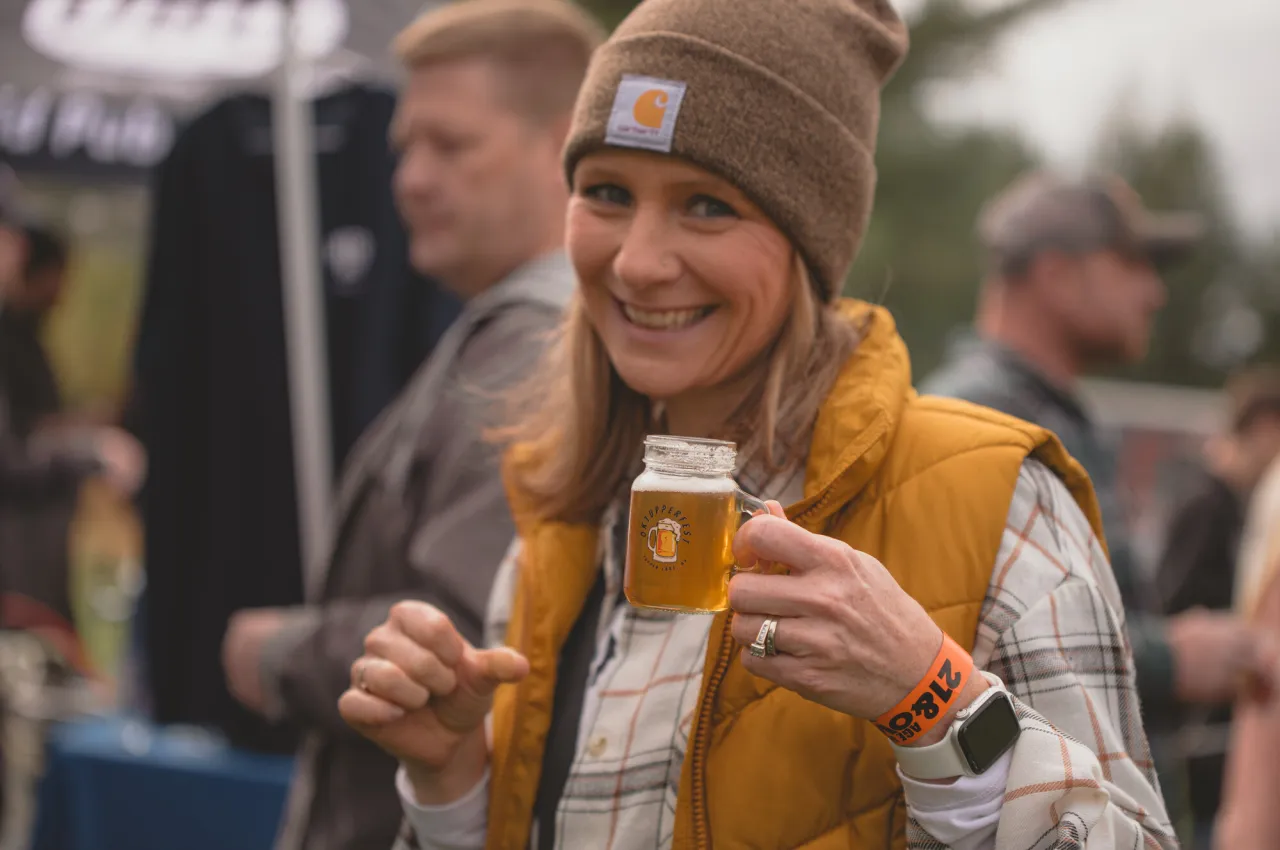A woman in a brown Carhartt beanie and Carhartt vest holds up a mini mug of beer.