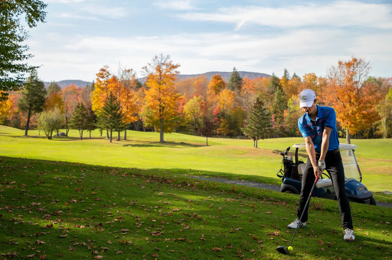 A golfer prepares to take a swing on a green with bright orange and yellow fall foliage in the forest beyond.