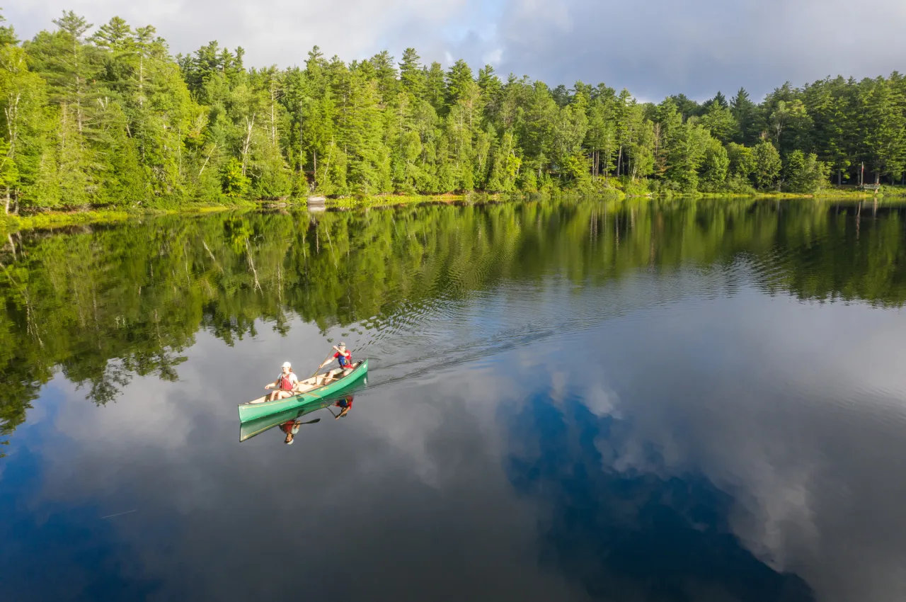 A canoe with two paddlers in it glides across a calm lake surrounded by trees on a sunny day.