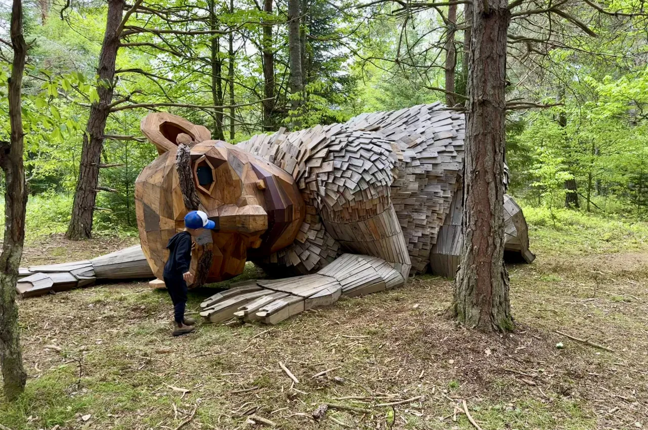 A toddler examines a gigantic wooden troll sculpture in a wooded forest.