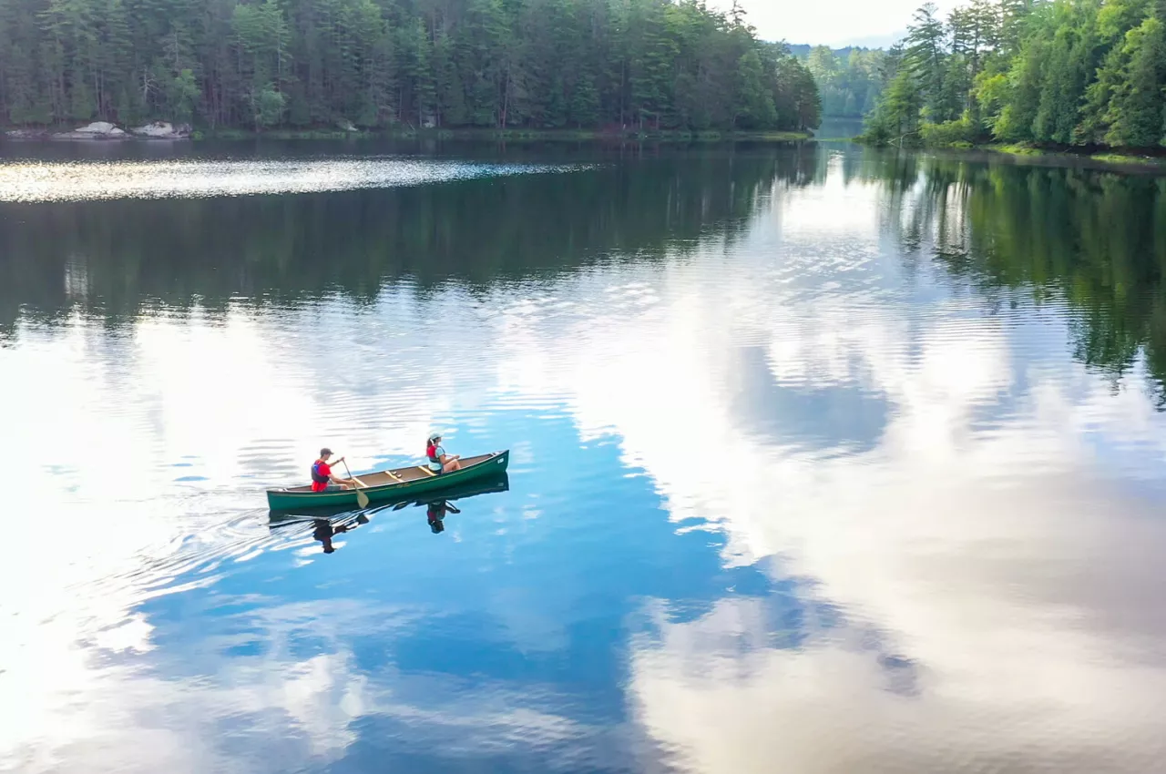 Two people paddle a canoe across an Adirondack lake that is reflecting the partly sunny sky