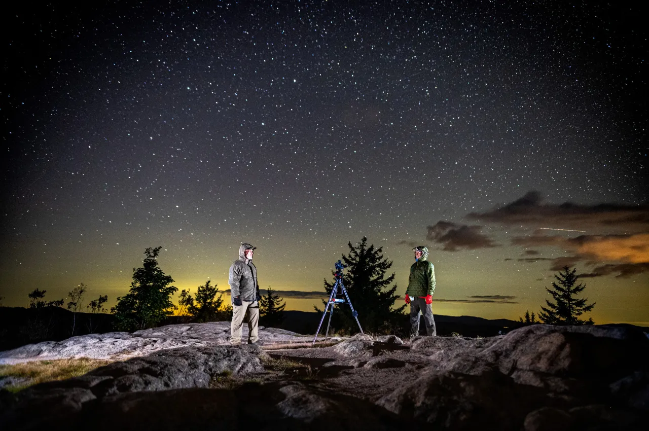 Two stargazers stand on top of a mountain summit with a starry sky above.