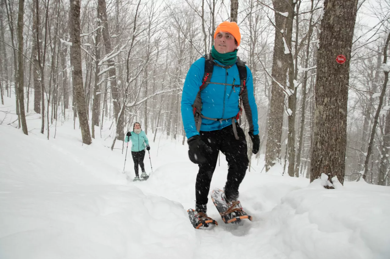 Two people snowshoe up an incline in a snowy woods