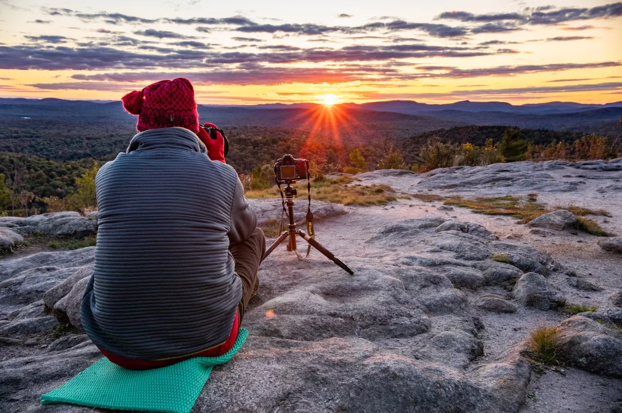 A photographer sits in front of a camera on a tripod atop a rocky summit at sunset.