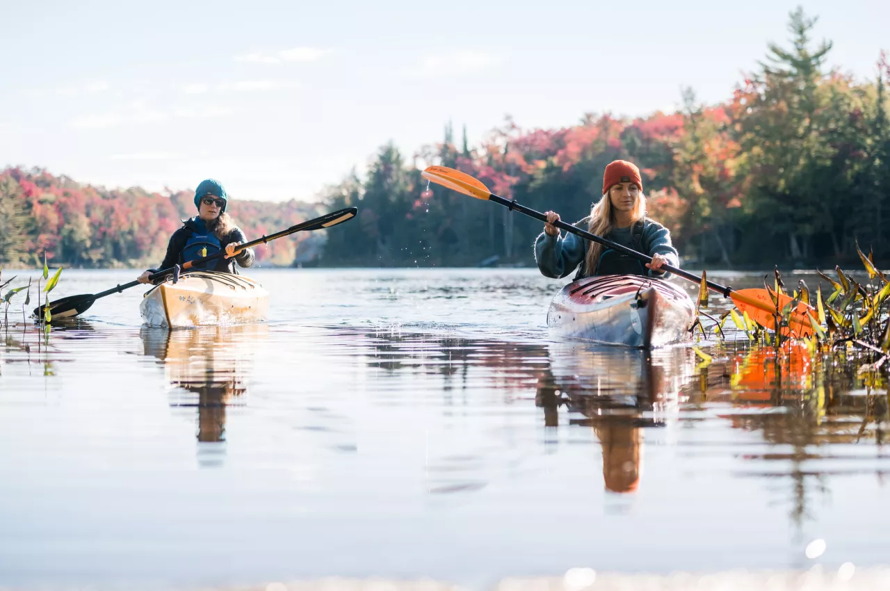 Two females paddle kayaks on a calm body of water with colorful fall foliage around them.
