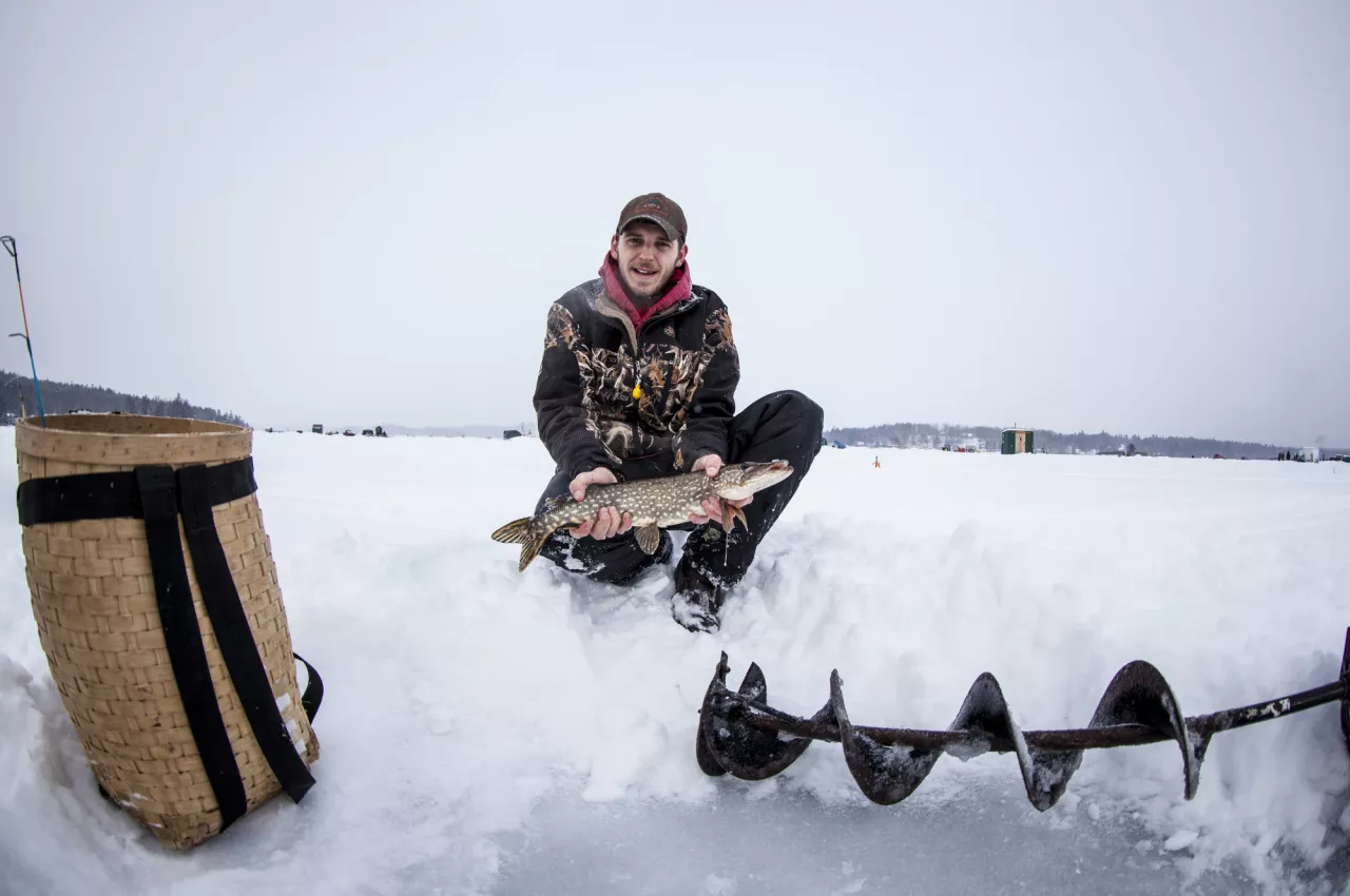 A man holds out a freshly caught fish on a field of ice, with an auger and packbasket beside him.