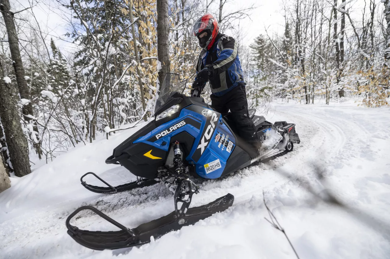 A snowmobiler stands up on a curve on a blue snowmobile in snowy woods.