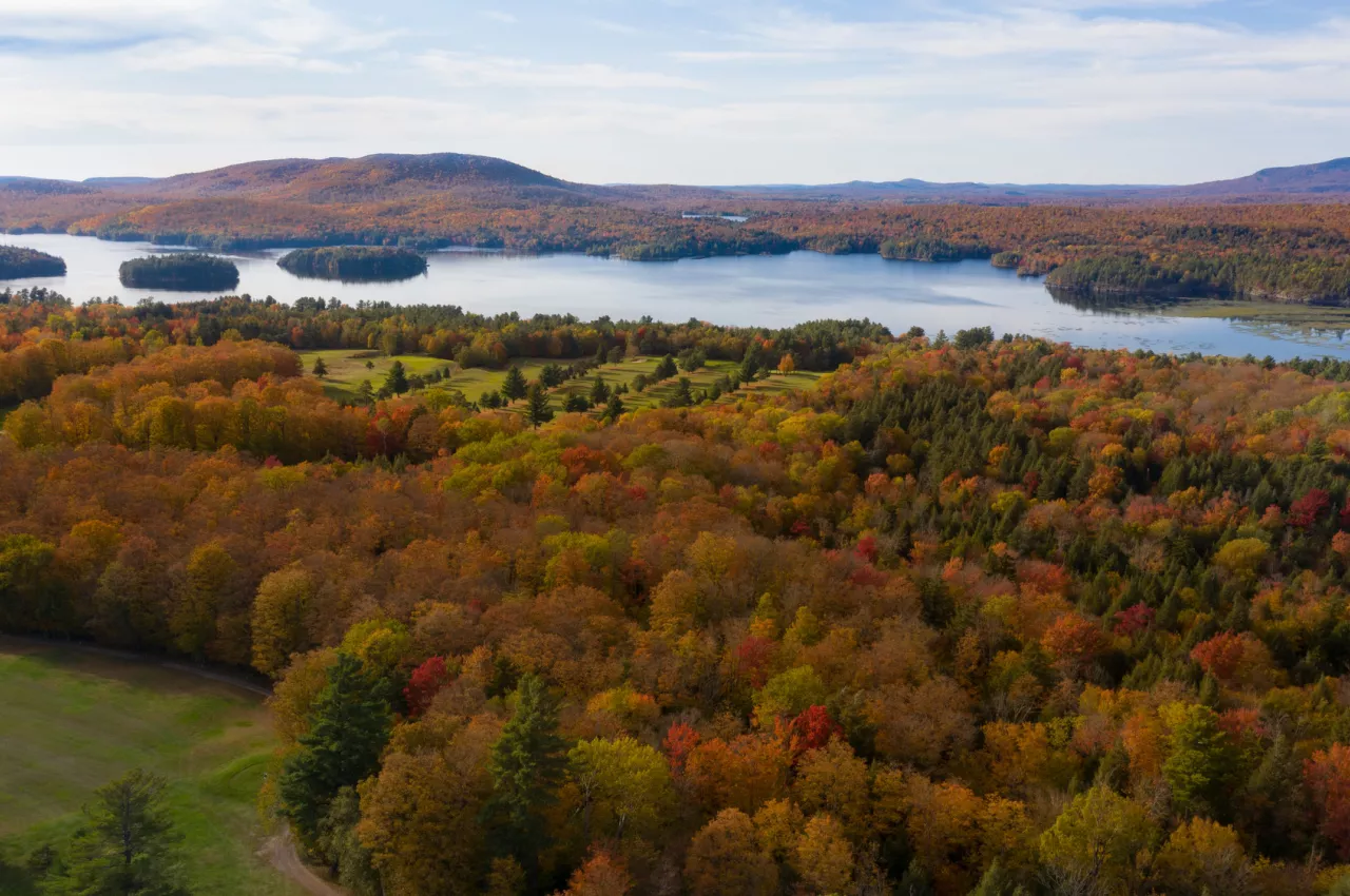 An aerial view of a brightly colored forest showing fall foliage, with a long, narrow lake in the distance.