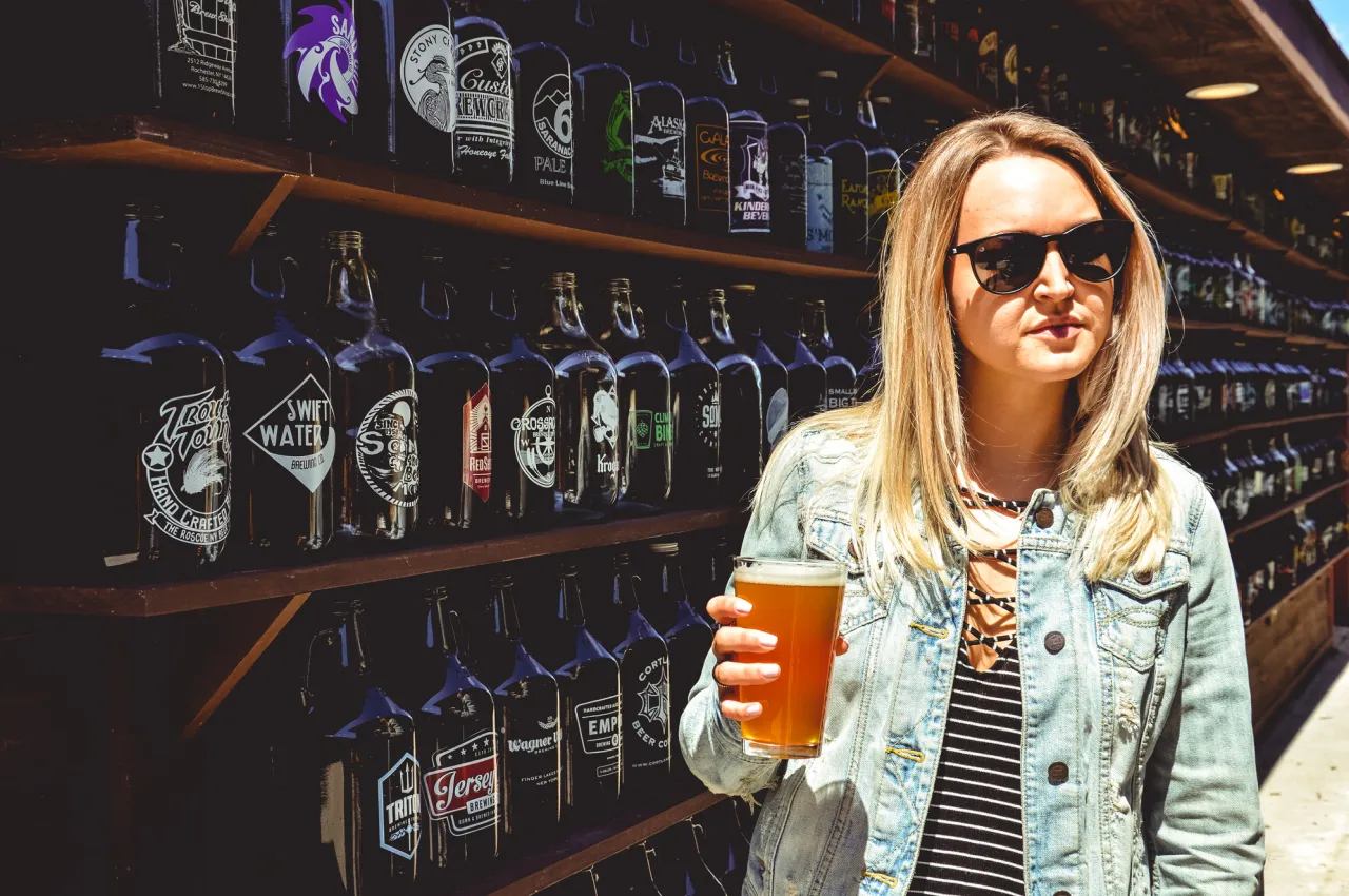 A blonde woman wearing sunglasses holds a glass of beer in front of a row of growlers.