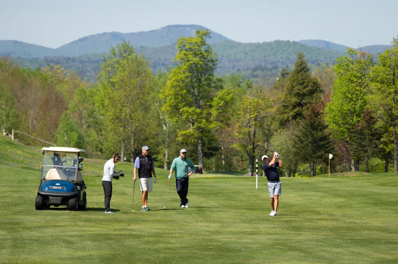 Three men watch while a fourth man tees off on a bright green golf course with mountains in the background.
