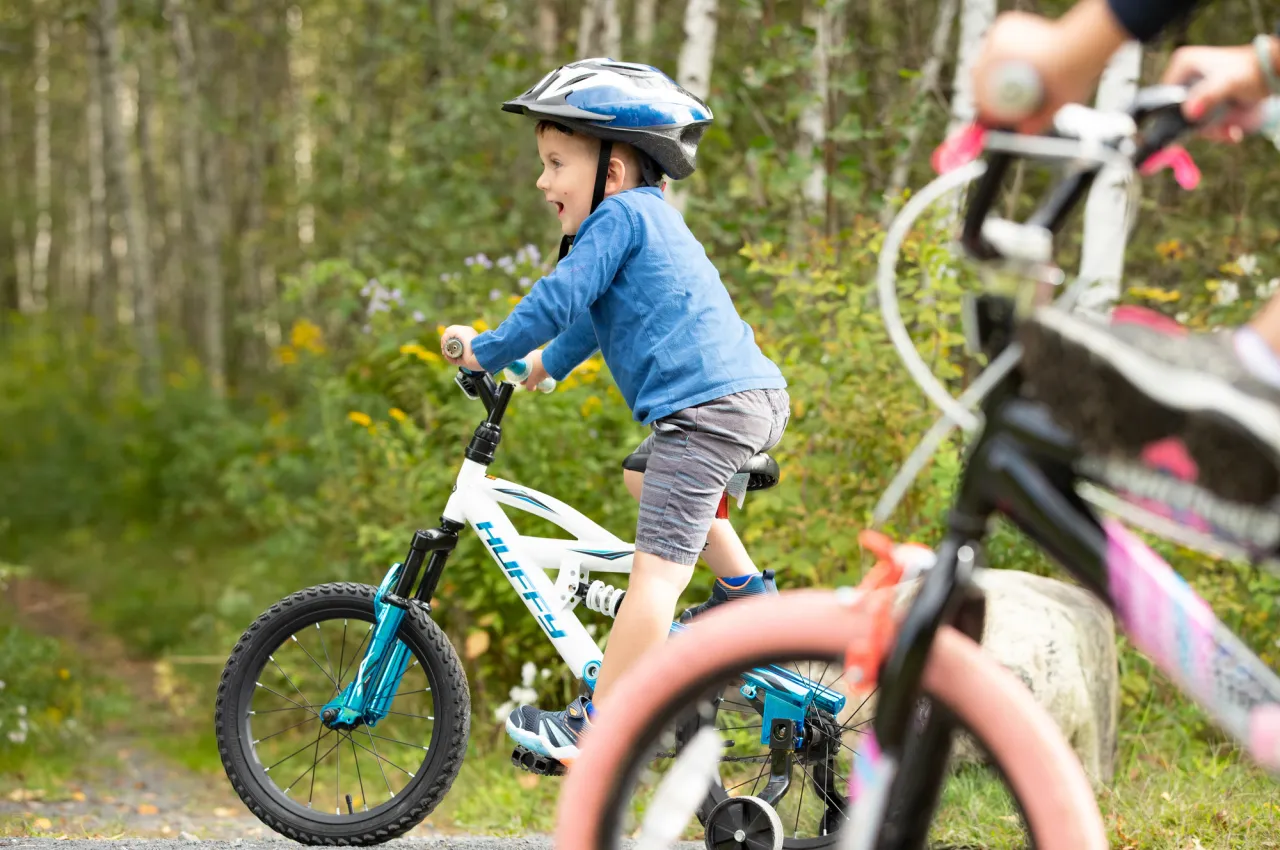 A young boy rides on a small bicycle, smiling, with trees in the background.