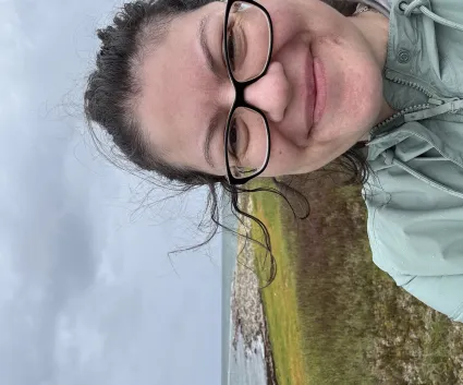 A woman with windblown hair smiles in front of a grey coastline.