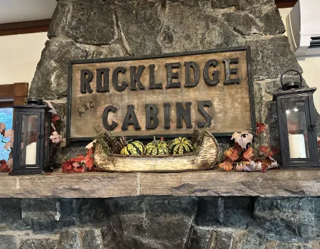 A carved sign reading 'Rockledge and Cabins' over a stone mantle with birch bark boat filled with festive squash and candles.