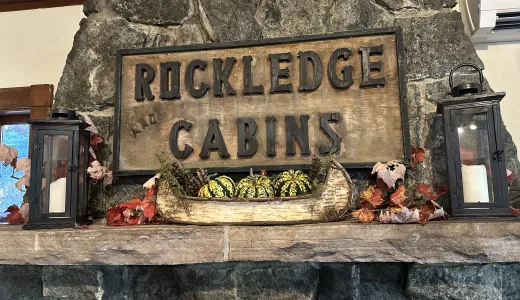 A carved sign reading 'Rockledge and Cabins' over a stone mantle with birch bark boat filled with festive squash and candles.