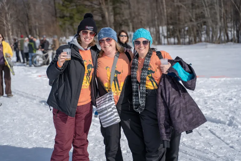 Three women grin and hold up cups of beer on a snowy field.