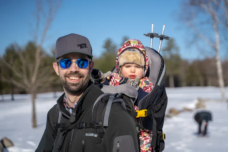 A man in sunglasses smiles with a baby in a pack on his pack on a sunny, snowy day.