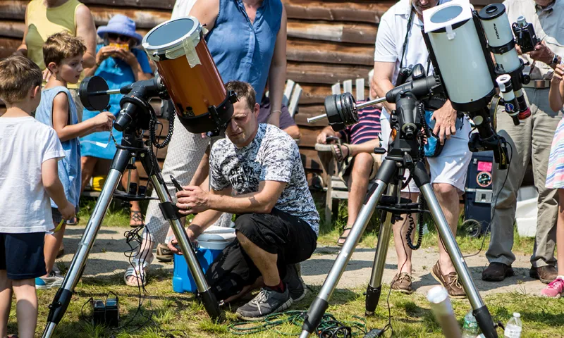 A man kneeling and using a telescope at an event with people around