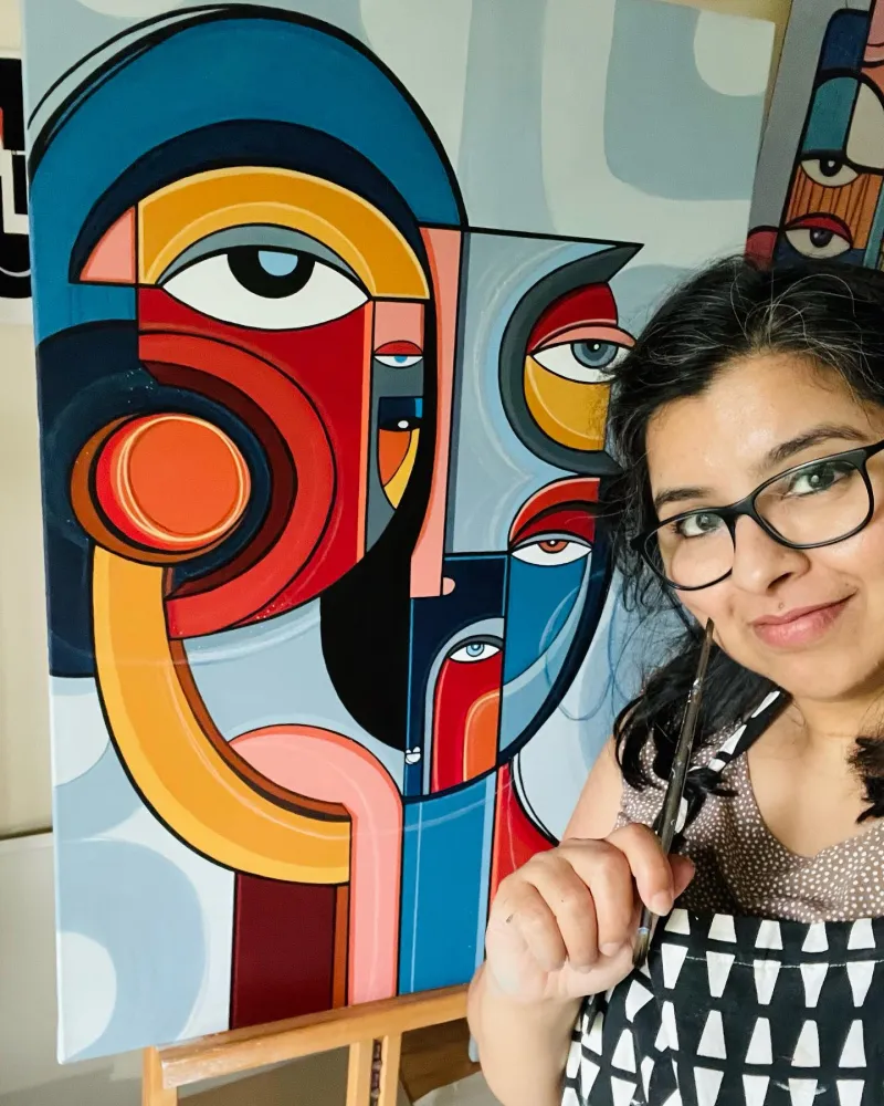A woman with dark hair and glasses poses next to a brightly colored cubist painting hanging on a wall.