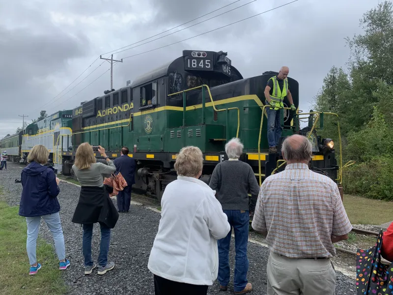 A group of people welcome a green and grey train into the Tupper Lake Station.
