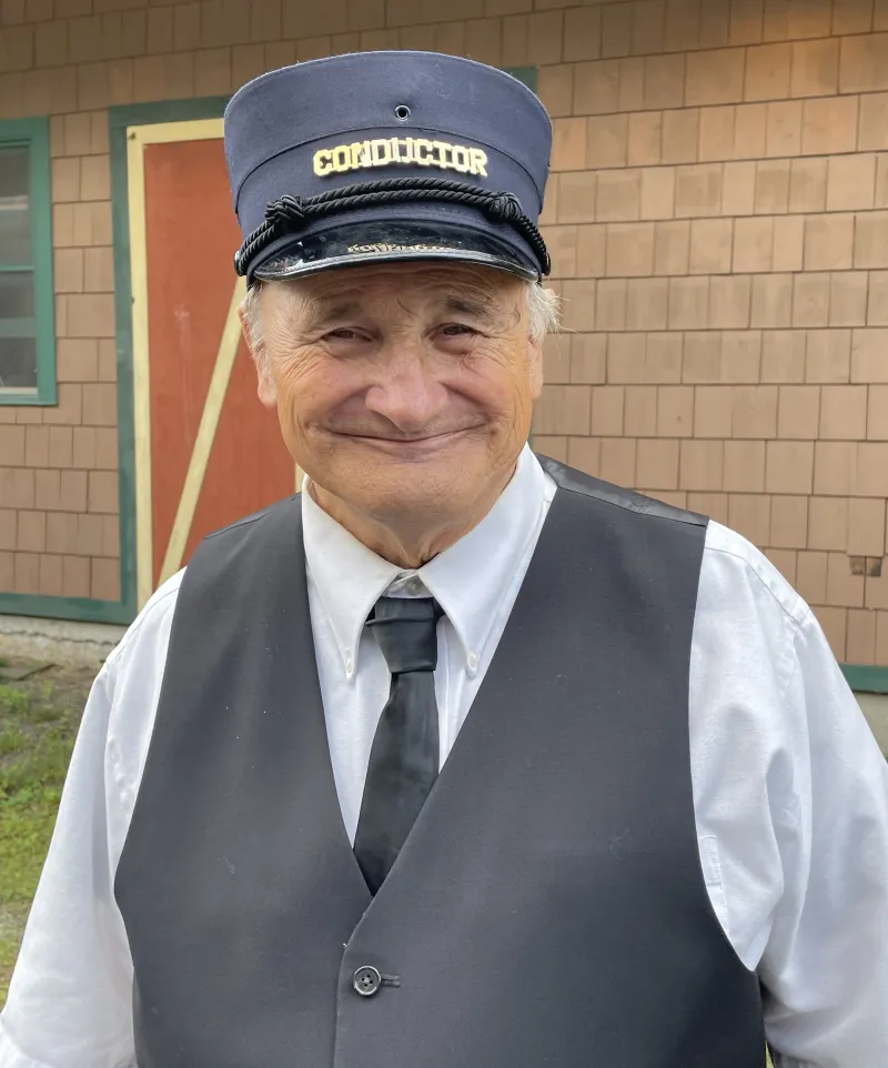 A grey-haired man in a vest, tie, and flat cap of a railroad conductor smiles at the camera.