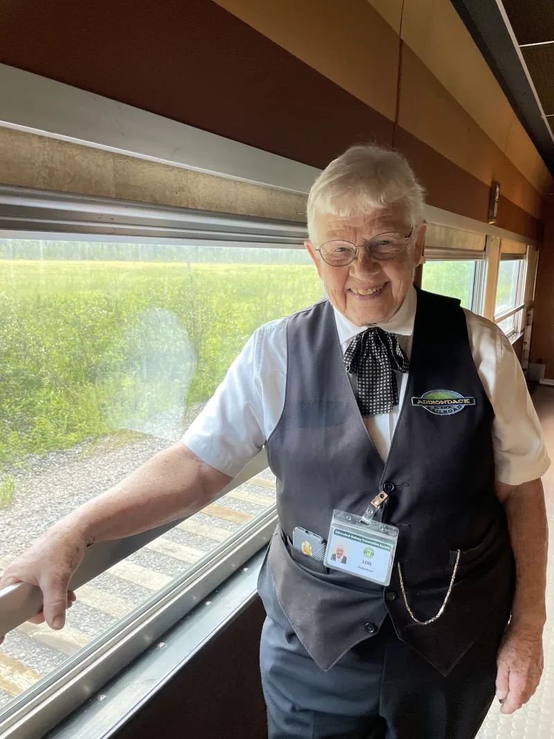 A woman with short white hair smiles happily in front of a window in a vintage railroad cafe car.
