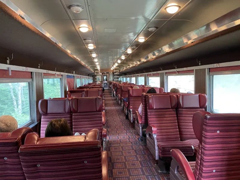 A view of the indoor cabin of a passenger train car, with red upholstered seats, carpeted aisle, and overhead lighting.