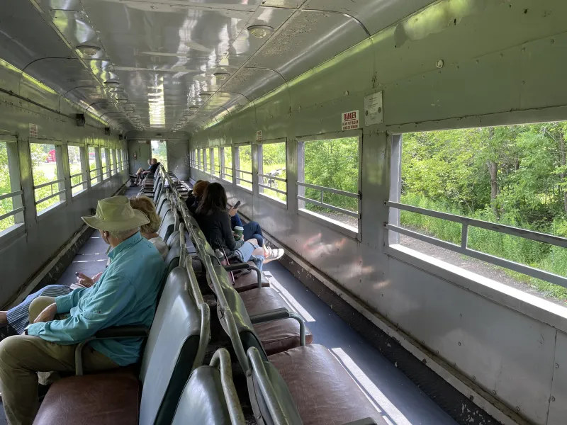 A view of the inside of a passenger open-air train car, with several people sitting in the seats looking out tr