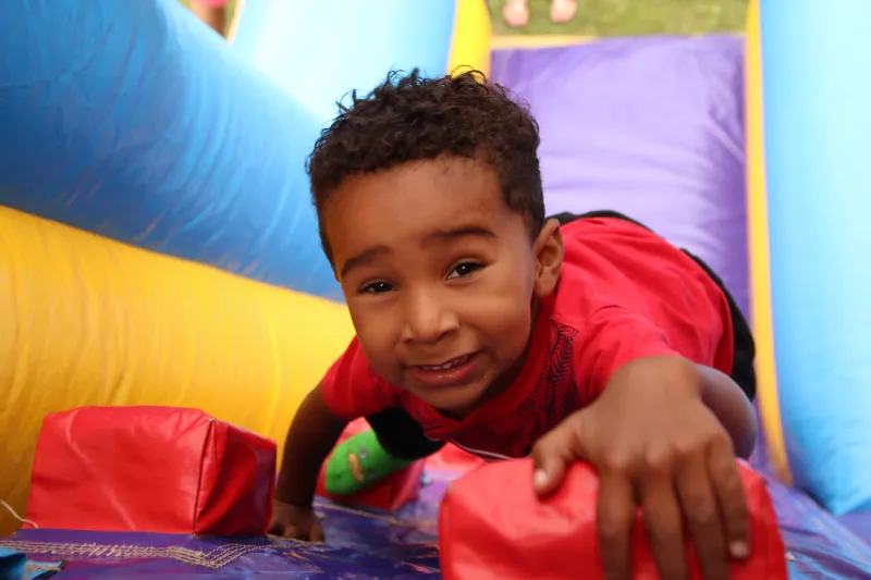 A small child smiles at the camera while climbing inside a colorful bounce house.