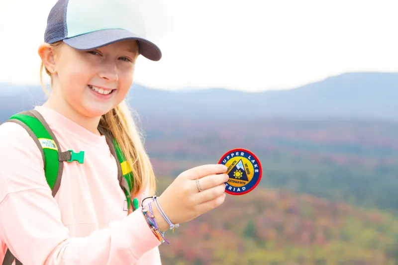 A teenage girl wearing a baseball cap poses on a mountain summit, holding a hiking challenge patch.
