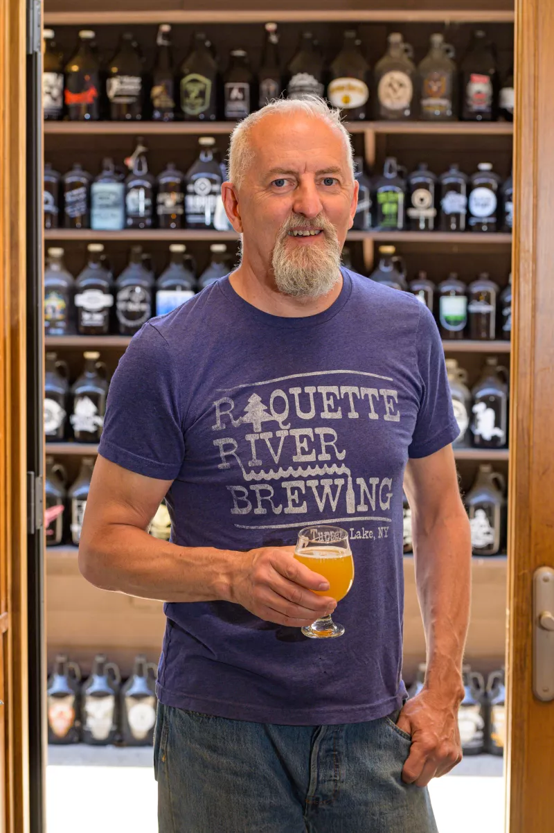 The co-owner of Raquette River Brewing, Mark Jessie, stands in front of growlers while holding a beer