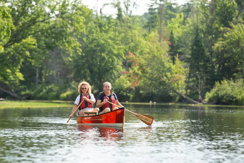 Two women paddle a red canoe along a river with trees in the background.
