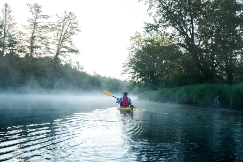 A solo kayaker paddling down a river in the morning fog.