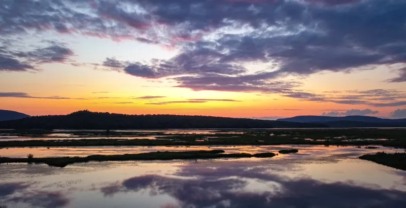 Sunset over Tupper Lake's wetlands with distant mountains and clouds.
