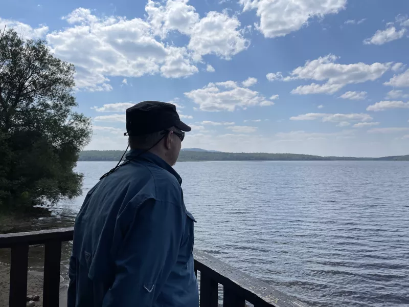 A man in a hat and sunglasses gazes at the view of a sparkling pond.
