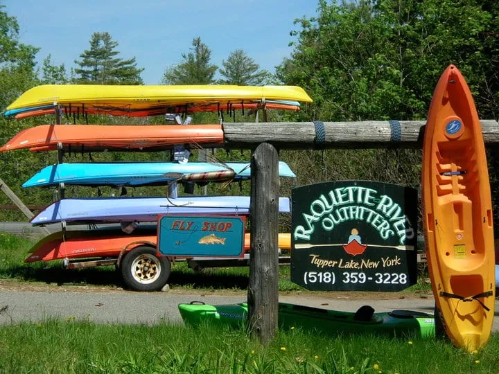 The business sign for Raquette River Outfitters with some of the rental kayaks on boat racks in the background