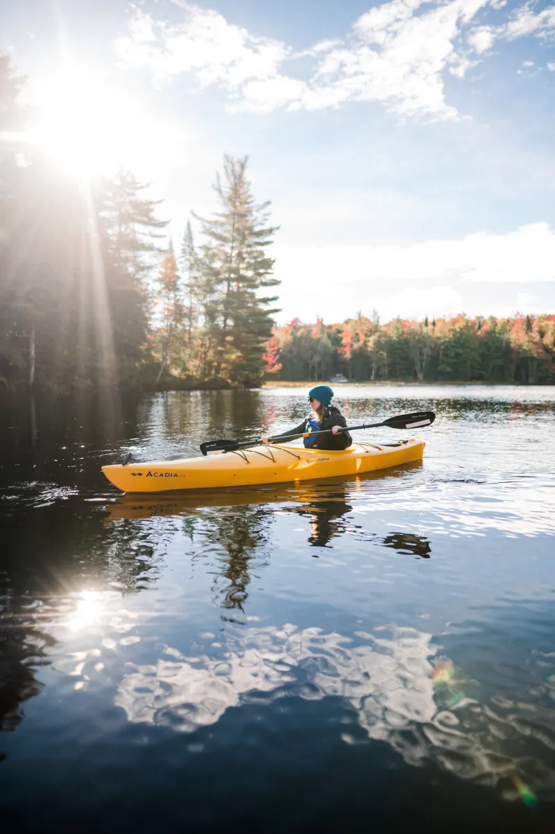 A woman paddles on a lake in a kayak with fall foliage along the shoreline.